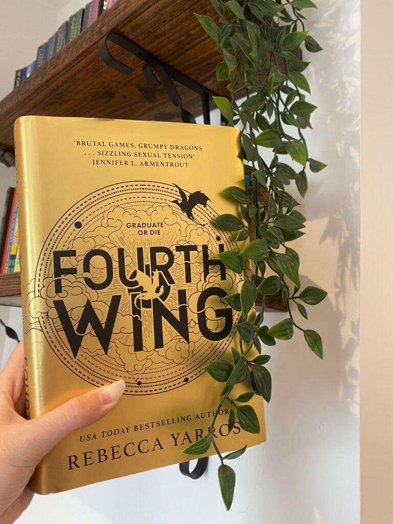 Fourth wing book held up next to a dark wood bookshelf and eucalyptus plant